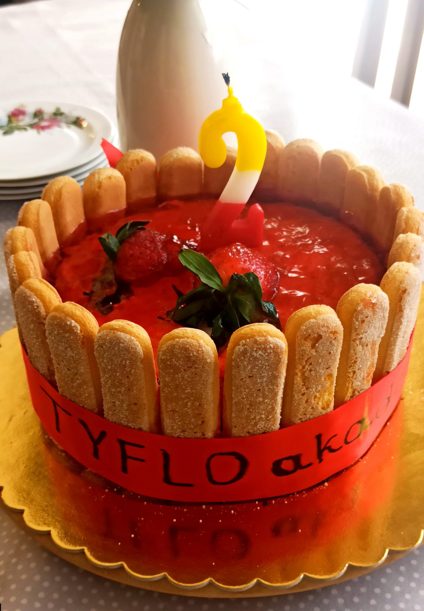 Tort urodzinowy TYFLOAkademi ze świeczką cyfry 2, przygotowany przez Basię. W tle wazon na kwiaty i talerzyki deserowe.