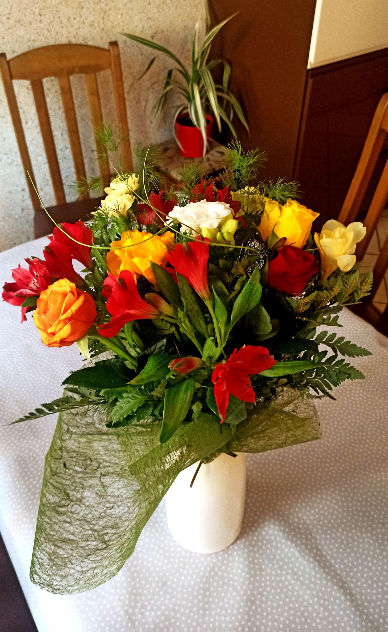 Kwiaty koloru czerwonego i żółtego z przybraniem w wazonie stojące na stole przyozdobionym białym obrusem, wokół stoją krzesła