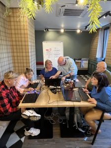 Na zdjęciu znajduje się grupa sześciu osób siedzących przy drewnianym stole, na którym znajdują się laptopy i różne kable. Osoby są zaangażowane w pracę na komputerach. Mężczyzna w jasnej bluzie pochyla się nad kobietą w niebieskiej bluzce pomagając jej w czymś na laptopie. W tle widać tablicę z informacjami, a na niej kilka logo, w tym logo z godłem Polski. Pomieszczenie ma nowoczesny wystrój z roślinami zwisającymi z sufitu i klimatyzacją. Podłoga jest wyłożona kafelkami w czarno-białą szachownicę.