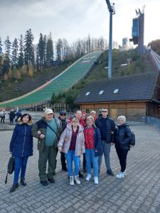 Na fotografii widzimy grupę dziesięciu osób stojących na placu wyłożonym kostką brukową. W tle znajduje się duża skocznia narciarska w Wiśle-Malince, wyróżniająca się zielonym zeskokiem oraz wieżą sędziowską po prawej stronie. Skocznia otoczona jest drzewami, częściowo ogołoconymi z liści, co wskazuje na jesienną aurę. Uczestnicy zdjęcia ubrani są w ciepłe stroje.