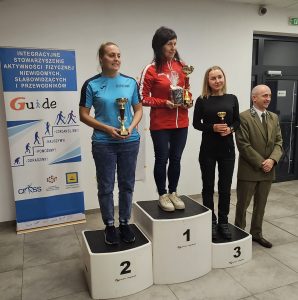 Na zdjęciu znajdują się trzy kobiety stojące na podium oraz mężczyzna w garniturze. Kobieta na najwyższym stopniu podium (oznaczonym numerem 1) ma na sobie czerwoną bluzę sportową z białymi paskami na rękawach i trzyma w rękach puchar oraz zapakowany prezent. Kobieta na drugim stopniu podium (oznaczonym numerem 2) ma niebieską koszulkę sportową z napisem "Jutrzenka" i również trzyma puchar. Kobieta na trzecim stopniu podium (oznaczonym numerem 3) ubrana jest na czarno i także trzyma puchar. Obok podium stoi mężczyzna w zielonym garniturze, który się uśmiecha. W tle znajduje się baner z napisem "Integracyjne Stowarzyszenie Aktywności Fizycznej Niewidomych, Słabowidzących i Przewodników Guide" oraz hasłami "Zorganizujemy!", "Nauczymy!", "Pomożemy!", "Doradzimy!". Baner zawiera także logotypy różnych organizacji.