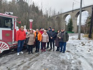 Na zdjęciu znajduje się grupa ludzi stojących na zaśnieżonej drodze. W tle widać czerwono-biały pojazd stylizowany na lokomotywę. Za grupą znajduje się wysoki wiadukt kolejowy z kilkoma łukami, a w tle widać drzewa bez liści, co sugeruje zimową porę roku. Osoby na zdjęciu są ciepło ubrane, noszą kurtki i czapki.