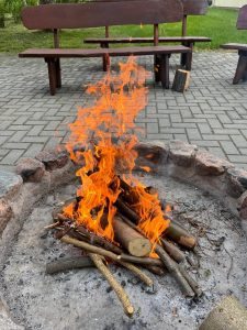 Ognisko Ognisko z jasnym, pomarańczowym płomieniem. Ogień pali się na stosie drewnianych gałęzi i kawałków drewna, ułożonych w kamiennym kręgu na ziemi. W tle widać dwie drewniane ławki ustawione na brukowanym placu, a za nimi trawnik. Na jednej z ławek stoi kawałek drewna.