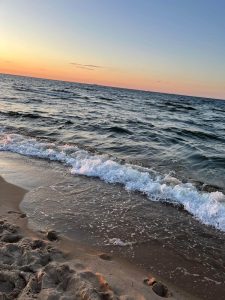 Plaża nad morzem o zachodzie słońca Na pierwszym planie widać mokry piasek z odciśniętymi śladami stóp oraz niewielkie fale rozbijające się o brzeg. Woda jest lekko wzburzona, a fale mają białą pianę. Na horyzoncie morze styka się z niebem, które jest rozświetlone ciepłymi barwami zachodzącego słońca, odcienie pomarańczowego, różowego i niebieskiego. Całość tworzy spokojny, letni klimat.