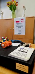 Biurko z różnymi materiałami edukacyjnymi Grupa osób w sali, w której kobieta w granatowym fartuchu medycznym tłumaczy coś, gestykulując. Na pierwszym planie siedzi mężczyzna tyłem do obiektywu, a na drewnianym stole leżą pomoce edukacyjne: maszyna do pisania brajlem, kostki Rubika, notatnik i inne. W tle stoi pięć kobiet, jedna z nich trzyma pomarańczowy przedmiot.