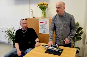 Instruktorzy podczas szkolenia Dwóch mężczyzn w pomieszczeniu. Jeden z nich siedzi przy stole, ma na sobie czarną koszulkę i okulary przeciwsłoneczne na głowie, drugi stoi obok stołu i pokazuje maszynę do pisania brajlem. W tle znajduje się drewniana szafka, na której stoi wazon z kolorowymi tulipanami oraz figurka anioła. Na szafce przyklejony jest plakat z informacją o dofinansowaniu ze środków Państwowego Funduszu Rehabilitacji Osób Niepełnosprawnych. Po bokach szafki stoją zielone rośliny doniczkowe.