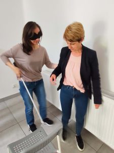 Wsparcie i zaufanie Ujęcie z góry przedstawia dwie kobiety w pomieszczeniu. Kobieta po lewej, z zawiązanymi oczami i białą laską, wyciąga rękę. Kobieta po prawej, prowadzi ją, trzymając jej ramię.