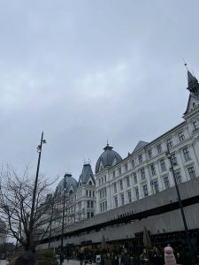 Zdjęcie przedstawia okazałą, białą kamienicę w stylu neorenesansowym pod pochmurnym, szarym niebem w Oslo. Budynek charakteryzuje się licznymi wieżyczkami z ciemnoszarymi, spiczastymi dachami i kopułami. Na dole widoczna jest nowoczesna, betonowa konstrukcja pasażu z napisem „OMAKASE”, pod którą znajdują się witryny sklepowe i kawiarnie.