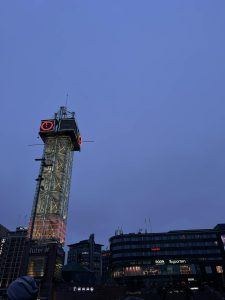 Nowoczesna, przeszklona wieża komunikacyjna Ruter# w centrum Oslo o zmierzchu. Wysoka konstrukcja z czerwonym podświetleniem i neonowym zegarem na szczycie dominuje nad placem. W tle widać rozświetlone budynki centrum handlowego Byporten i hotele pod granatowym niebem.